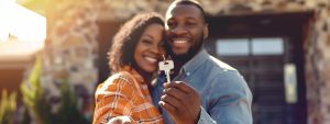 Couple smiling and holding house keys in front of a new home, symbolizing homeownership and real estate success in North Carolina.
